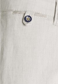 Světle šedá denimmová látka s viditelným švem, s kulatým modrým knoflíkem se žlutým středem a malým vrchním otvorem na opasek.