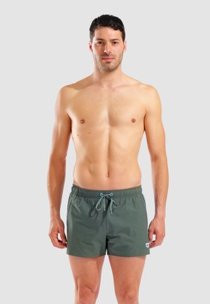 Mann mit kurzen dunklen Haaren, der grüne Badeshorts mit Kordelzug trägt und vor einem schlichten hellen Hintergrund steht.