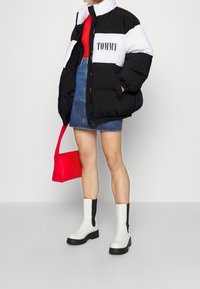 Giacca a vento nera e bianca con logo "TOMMY", abbinata a una gonna di jeans e una borsa rossa. Stivali alla caviglia bianchi con dettagli neri.