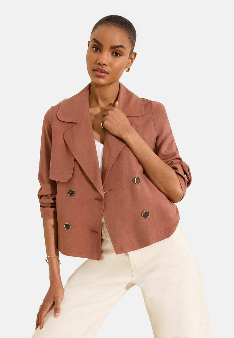 Young woman wearing a rust-colored double-breasted blazer with rolled sleeves and off-white pants, posing against a plain background.