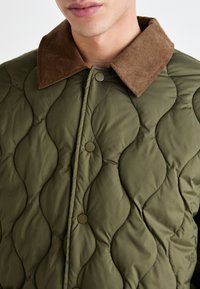 Quilted olive green jacket with a brown corduroy collar, featuring snap buttons and a wavy texture pattern across the fabric.