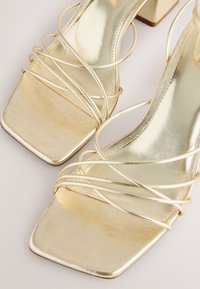 Sandales dorées à brides avec bouts carrés et petits talons blocs sur un fond clair.