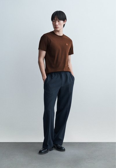 Homme debout les mains dans les poches, portant une chemise marron à manches courtes, un pantalon ample foncé et des chaussures noires, sur un fond clair uni.