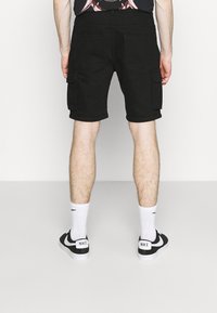 Černé cargo šortky se bočními kapsami, ohrnutým lemem a rovný střih. Nosí se s bílými ponožkami a černými teniskami Nike. Viditelná kůže na nohách.