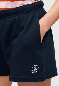 Σκούρα μπλε σορτς με ελαστική μέση, πλαϊνή τσέπη, λευκό κεντημένο λογότυπο "FH" κοντά στο τελείωμα, φορεμένα με το χέρι στην τσέπη και ορατό ριγέ πορτοκαλί πουκάμισο.