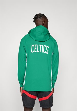 Osoba s pletenými vlasmi, oblečená v zelenej mikine s kapucňou s nápisom „CELTICS“ na chrbte a čiernych kraťasoch s červenými pruhmi, stojaca chrbtom.