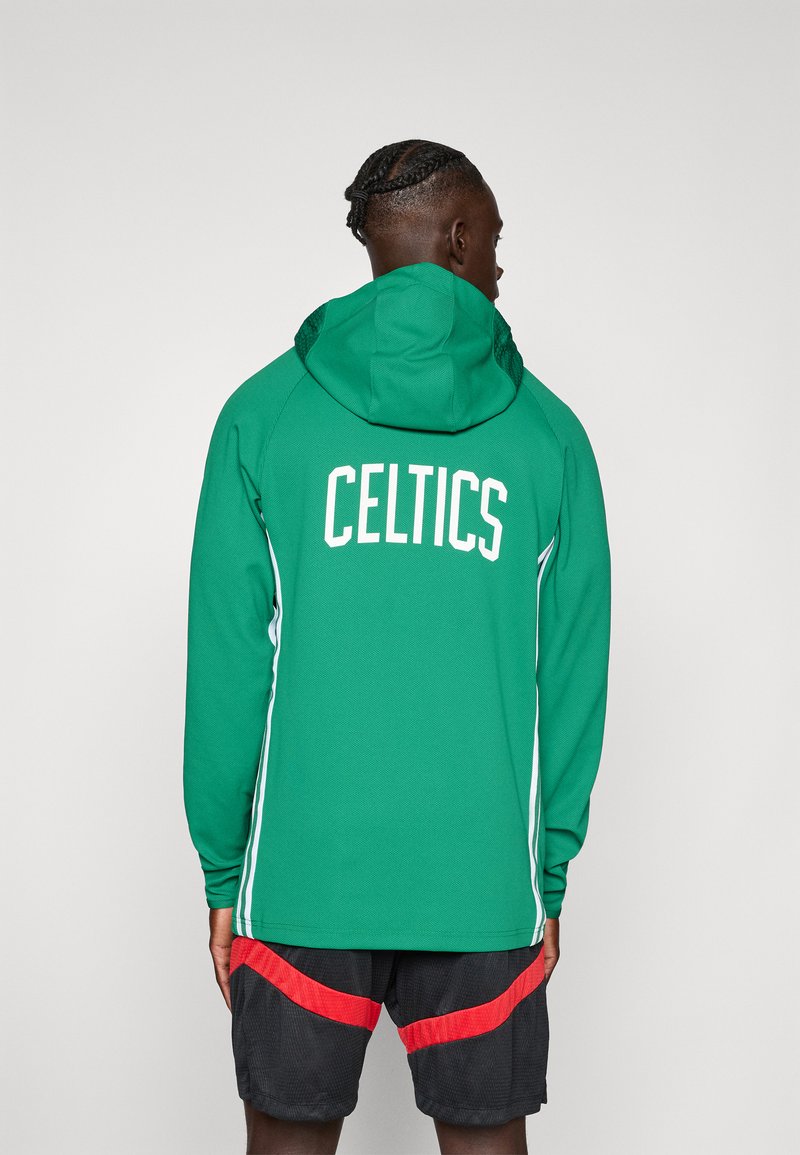 Punutud juustega inimene, kes kannab rohelise kapuutsiga dressipluusi, millel on taga kiri "CELTICS", ning musti lühikesi pükse punaste triipudega, seisab seljaga vaataja poole.