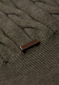 Zelený pletený svetr s cable-knit vzorem, který má obdélníkovou kovovou značku s vyraženým nápisem "QUINTESSENCE".