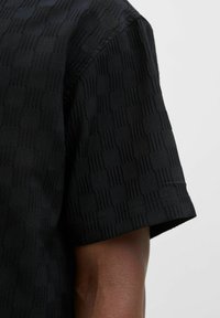Chemise noire à manches courtes avec un motif texturé et des dessins rectangulaires en relief, fabriquée dans un tissu doux avec une finition lisse.