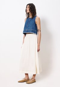 Denim-Cropped-Top mit rohem Saum, kombiniert mit einem luftigen, plissierten weißen Maxikleid. Beige Schnürschuhe runden das Outfit ab.