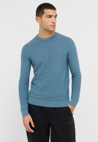 Maglione blu a coste con collo rotondo e maniche lunghe. Presenta un piccolo logo sul petto. Abbinato a pantaloni scuri.