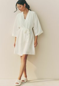 Biały szlafrok w stylu kimono z szerokimi rękawami, ściągnięty w talii sznurkiem. W zestawie z przytulnymi białymi kapciami z dekoracyjnymi akcentami.