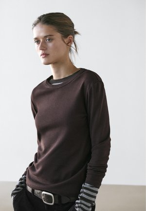 Ung kvinde med opsat hår iført en mørkebrun langærmet trøje, stripede handsker og sorte bukser med bælte, stående mod en ensfarvet baggrund.