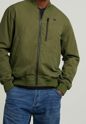 Homme portant une veste bomber vert olive avec zip et poches latérales, et un jean bleu, les mains dans les poches, sur fond uni.