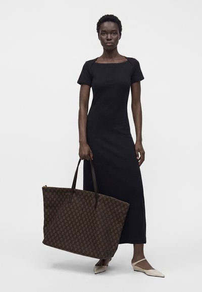 Femme portant une robe noire à manches courtes, longueur cheville, tenant un grand sac cabas marron à motifs, debout sur un fond blanc.