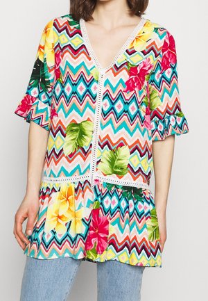 Woman wearing a colorful zigzag patterned blouse with large tropical flowers and ruffled short sleeves, paired with light blue jeans.