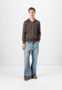 Pulover cu fermoar de culoare maro închis, blugi decolorati lejeri de culoare albastră deschisă și pantofi slip-on maro. Vestimentația are un stil relaxat și confortabil.