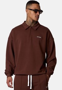 Pull en brun avec un col zippé, fabriqué en tissu doux. Présente un logo sur la poitrine et des poignets côtelés. Assorti avec un pantalon coordonné.