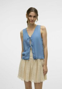 Blå ärmlös blouse med V-ringning och knytning, tillsammans med en beige spetskjol med en scalloped mönster.