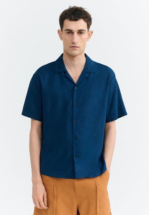 Junger Mann, der ein marineblaues kurzärmeliges Hemd mit Knöpfen und senffarbene Hose trägt, steht vor einem einfachen weißen Hintergrund.