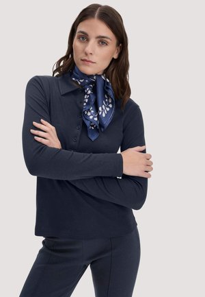 Frau mit braunen Haaren, die ein marineblaues Poloshirt mit langen Ärmeln, passende Hosen und einen marineblauen Schal mit weißen Blattmustern trägt, die Arme verschränkt.