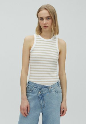 Jeune femme aux cheveux blonds mi-longs, portant un haut sans manches blanc et beige rayé et un jean bleu taille haute.