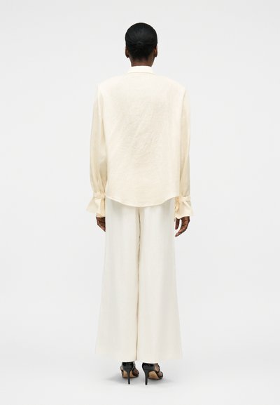 Άτομο με σκουρόχρωμα μαλλιά σε κότσο, που φοράει εκρού μακρυμάνικο μπλουζάκι με κορδόνια στους καρπούς, ταιριαστό παντελόνι με φαρδιά μπατζάκια και μαύρα ψηλοτάκουνα σανδάλια.