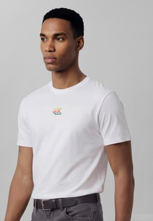 Νέος άνδρας που φοράει λευκό μπλουζάκι με μικρό πολύχρωμο λογότυπο "cK" στο στήθος, σκούρο ζωνάρι και γκρι παντελόνι, στέκεται μπροστά σε απλό γκρι φόντο.