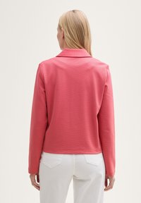 Veste longue à manches longues de couleur rose corail avec un col, en tissu texturé et coupe droite, accompagnée de pantalons blancs. La vue arrière montre l'ourlet et les manches.