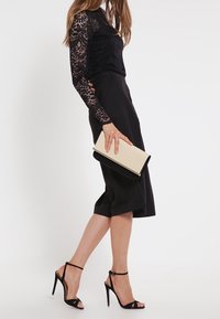 Top lungo a maniche nere in pizzo abbinato a una gonna nera al ginocchio, tacchi alti neri con cinturini e una clutch beige con dettagli neri.