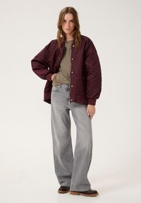 Maroon bomber jacket with snap buttons, olive green t-shirt, light gray wide-leg jeans, and dark brown shoes. Smooth fabric and relaxed fit.