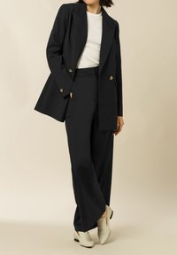 Blazer sartoriale blu navy scuro con doppia abbottonatura, abbinato a pantaloni a gamba larga coordinati. Maglietta bianca sotto e scarpe di colore chiaro.