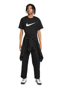 T-shirt Nike nera a maniche corte con logo swoosh bianco, abbinata a pantaloni larghi neri e una giacca nera annodata. Sneakers bianche e nere.