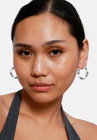 Silberne Creolen mit einer strukturierten Oberfläche, die eine kreisförmige Form haben. Das Modell trägt ein graues, gestreiftes Halstops. Glatte Haut und natürliches Make-up.