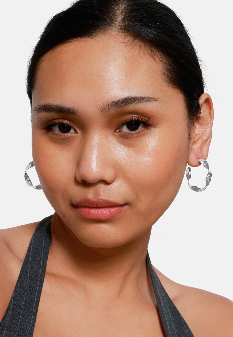 Silberne Creolen mit einer strukturierten Oberfläche, die eine kreisförmige Form haben. Das Modell trägt ein graues, gestreiftes Halstops. Glatte Haut und natürliches Make-up.