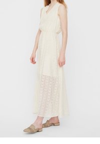 Langes, cremefarbenes Kleid mit ärmellosem Design und V-Ausschnitt. Verfügt über eine transparente untere Lage mit einem subtilen geometrischen Muster.