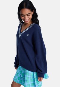 Πλεκτό πουλόβερ σε ναυτικό μπλε με λαιμόκοψη V και λευκές λωρίδες, συνδυασμένο με φούστα με πολλαπλά στρώματα, σε πράσινο και μπλε καρώ με βολάν.