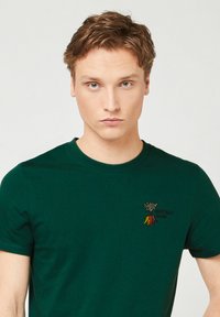 Jongeman met lichtbruin haar, die een donkergroen T-shirt draagt met een geborduurd kleurrijk wortelontwerp en de tekst "Vegetally Yours" op de borst.