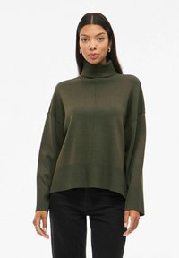 Pull à col roulé surdimensionné en vert olive avec des poignets et un ourlet côtelés. Texture en tricot douce et épaules tombantes. Associé à un pantalon noir.