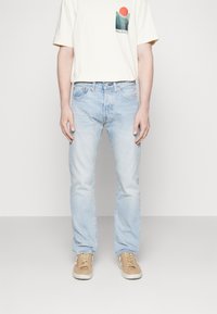 Jeans in denim azzurro con vestibilità dritta, abbinati a una T-shirt color crema con un design grafico verde e rosso. Sneakers beige.