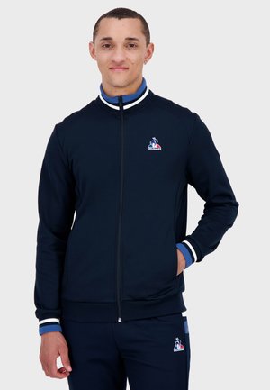 Navy zip-up jacket with white and blue striped cuffs. Features a logo on the left chest. Made of smooth fabric with fitted design.