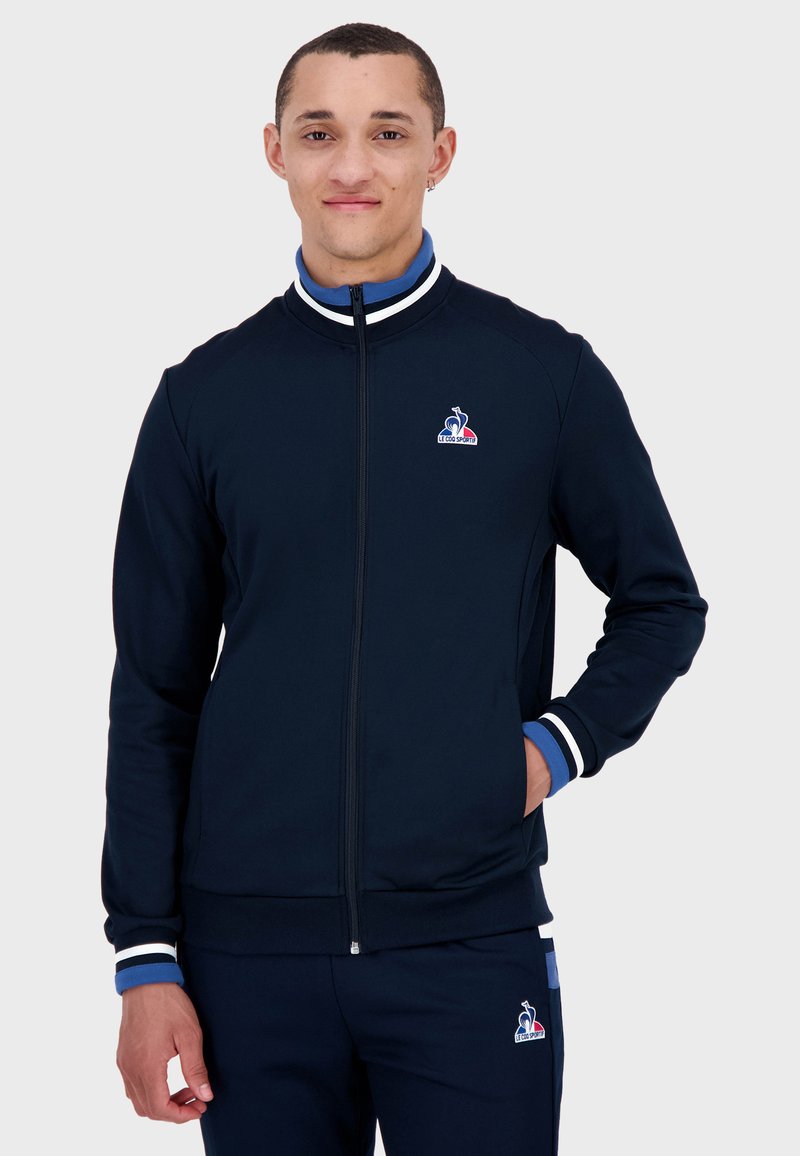 Giacca a zip blu navy con polsini a strisce bianche e blu. Presenta un logo sul lato sinistro del petto. Realizzata in tessuto liscio con un design aderente.