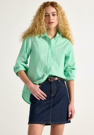 Grön- och vitrandig skjorta med knappar, ärmarna uppkavlade, kombinerad med en mörk jeanskjol och ett brunt bälte. Har en avslappnad passform och standardkrage.