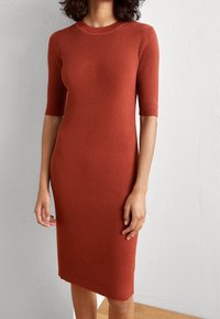 Robe en maille côtelée couleur orange brûlé avec un col rond et des manches courtes, présentant une silhouette ajustée qui s'affine légèrement jusqu'en dessous du genou.