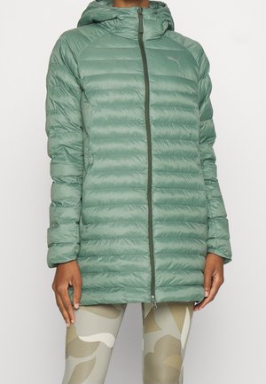 Personne portant une veste matelassée à capuche vert clair avec une fermeture éclair à l'avant et des leggings beiges à motifs.