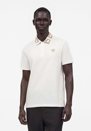 Homme portant un polo blanc à manches courtes avec un motif géométrique doré sur le col et un logo Medusa doré sur la poitrine, assorti à un pantalon foncé.