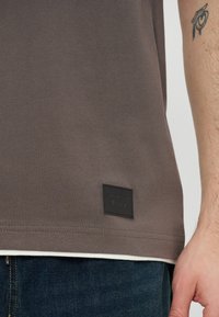 Gros plan sur le torse d'une personne portant une chemise marron avec un petit patch noir près de l'ourlet et un tatouage visible sur le bras droit.