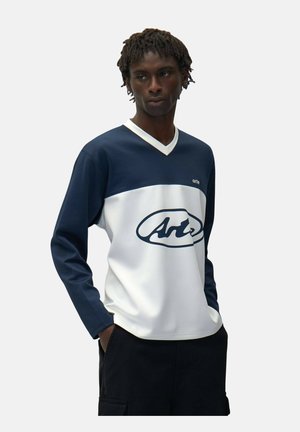 Giovane uomo dalla pelle scura e dreadlocks corte che indossa una maglietta a maniche lunghe blu navy e bianca con il logo "Arte", mani nelle tasche dei pantaloni neri.