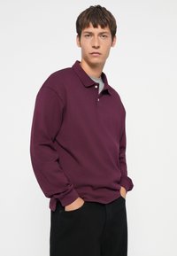 Maroon long-sleeve polo with a collar, featuring a three-button placket, worn over a gray T-shirt, paired with black pants. Smooth texture.