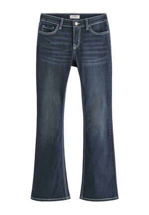 Ung person stående med hånden i lommen iført hvid kortærmet skjorte, mørkeblå jeans og hvide sneakers mod en ensfarvet baggrund.